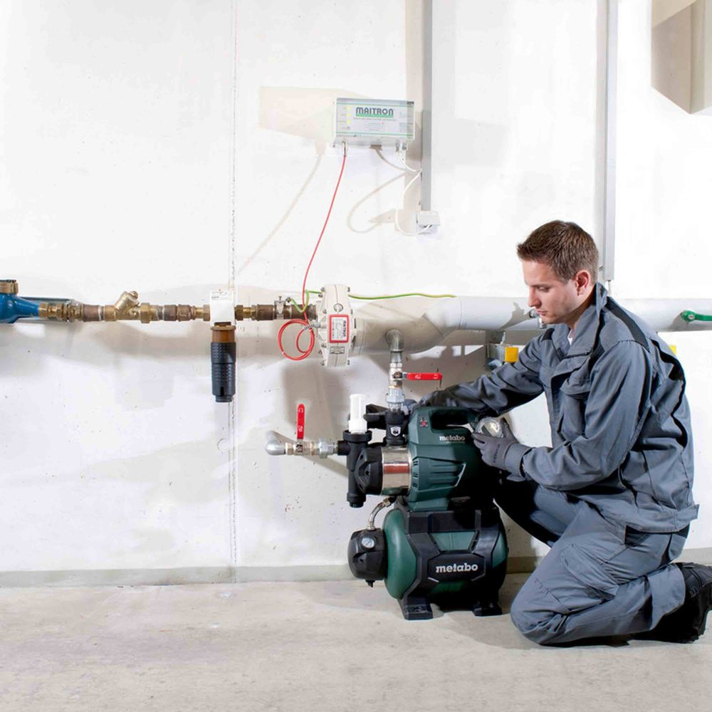 Mężczyzna w ubraniu roboczym klęczy, sprawdzając pompę wodną obok rur i urządzeń na ścianie.