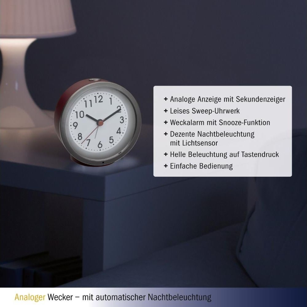Ein analoger Wecker mit weißem Zifferblatt und roten Zeigern steht auf einer dunklen Oberfläche.