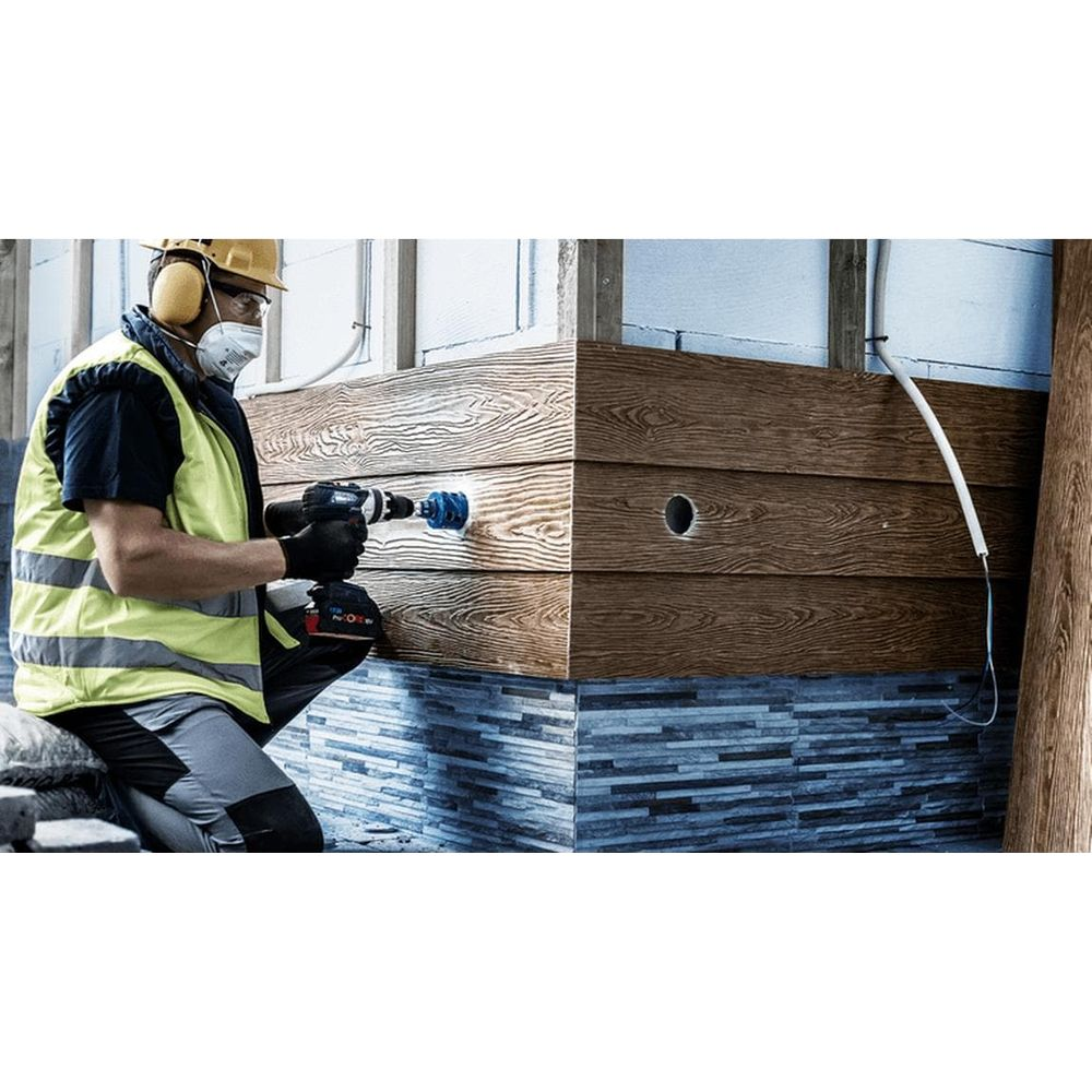 Mężczyzna wierci otwór w drewnianej elewacji, nosi sprzęt ochronny.