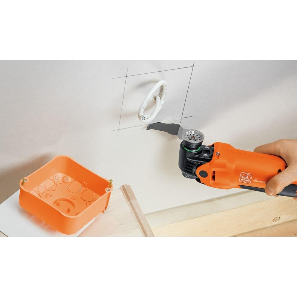 Ein orangefarbenes Multifunktionswerkzeug schneidet in eine weiße Wand neben einer orangen Einbaudose.