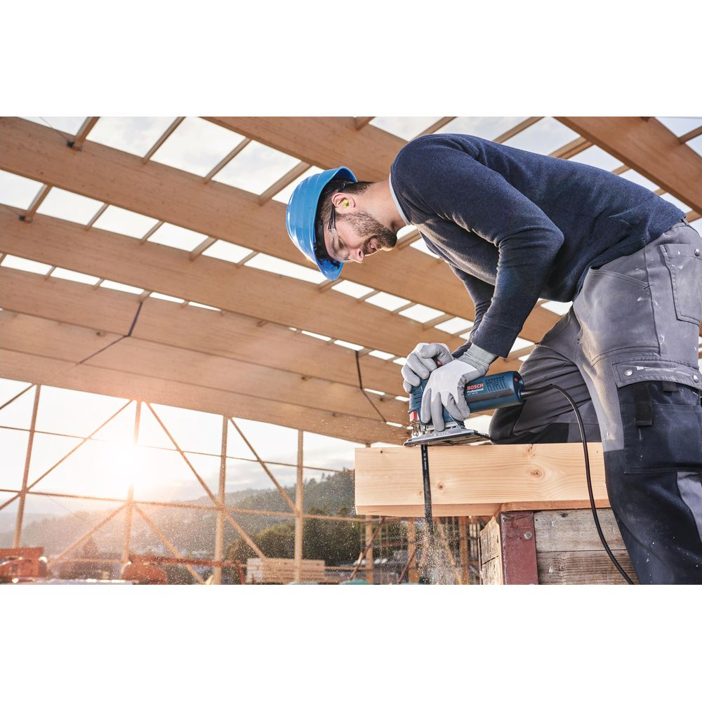 Robotnik w niebieskim kasku piłuje drewno, unosi się pył.