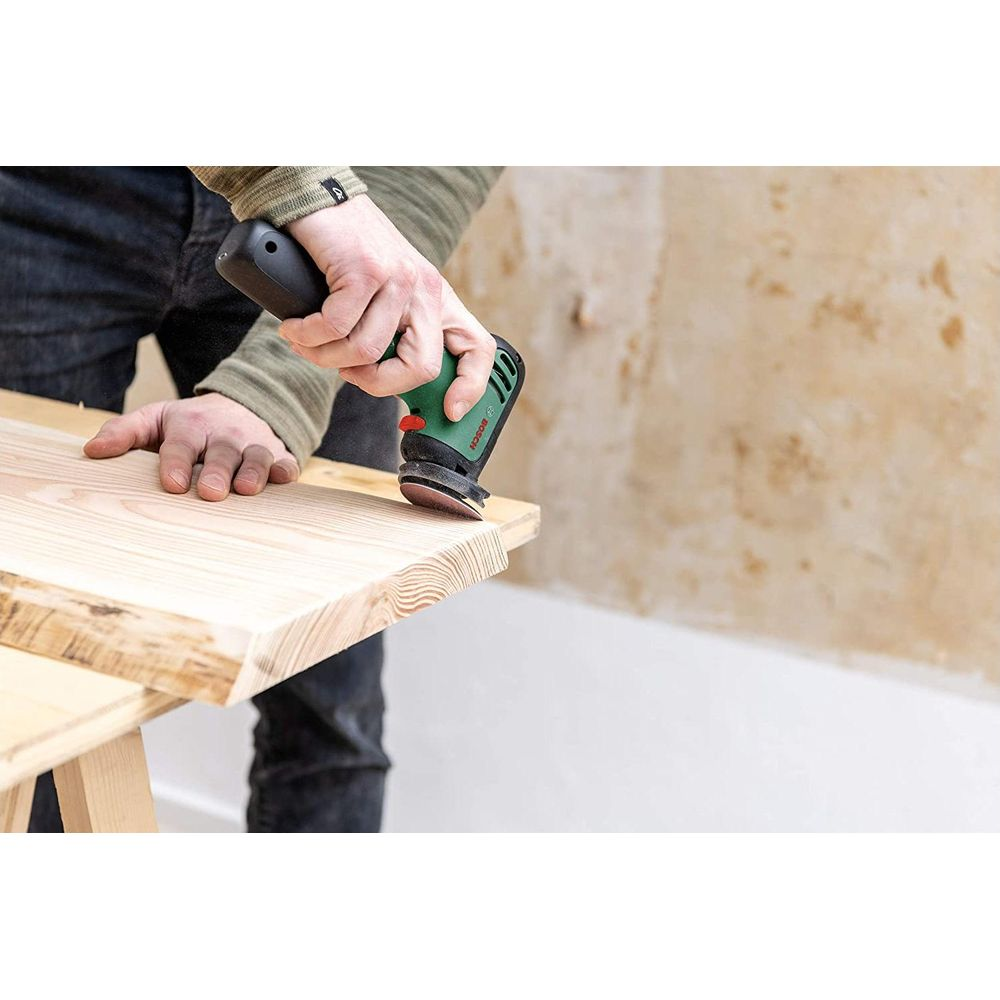 Eine Person schleift Holz mit einem grünen Bosch-Schleifgerät auf einem Holztisch.