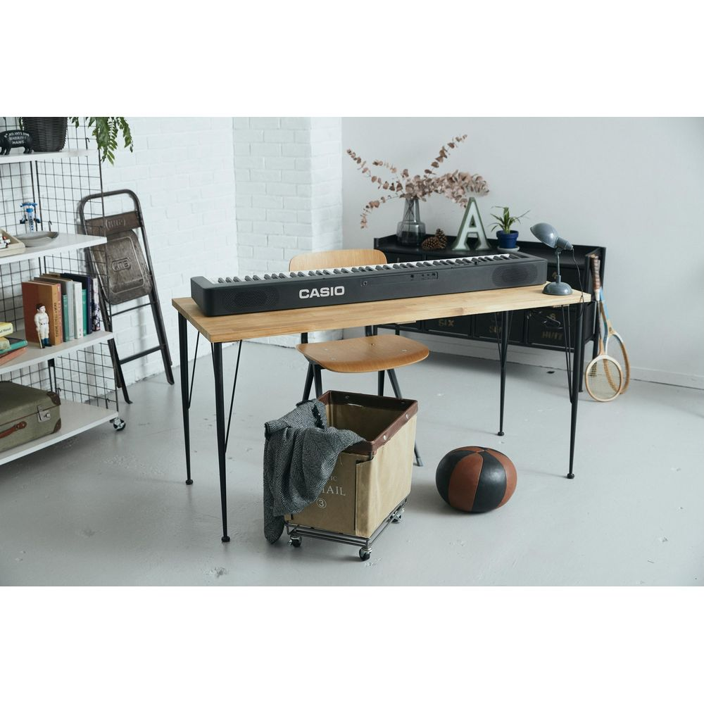 Ein schwarzes Keyboard steht auf einem Holztisch mit Metallbeinen. daneben ein Korb mit Decke.