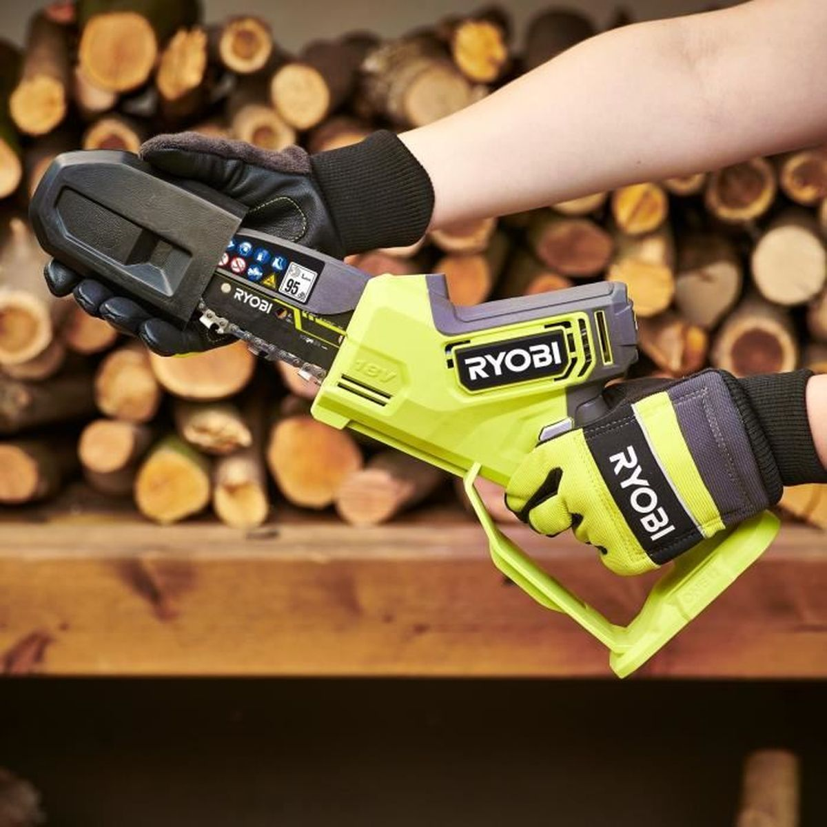 Hände in Ryobi-Handschuhen halten eine gelbe Kettensäge vor Holzstapeln.