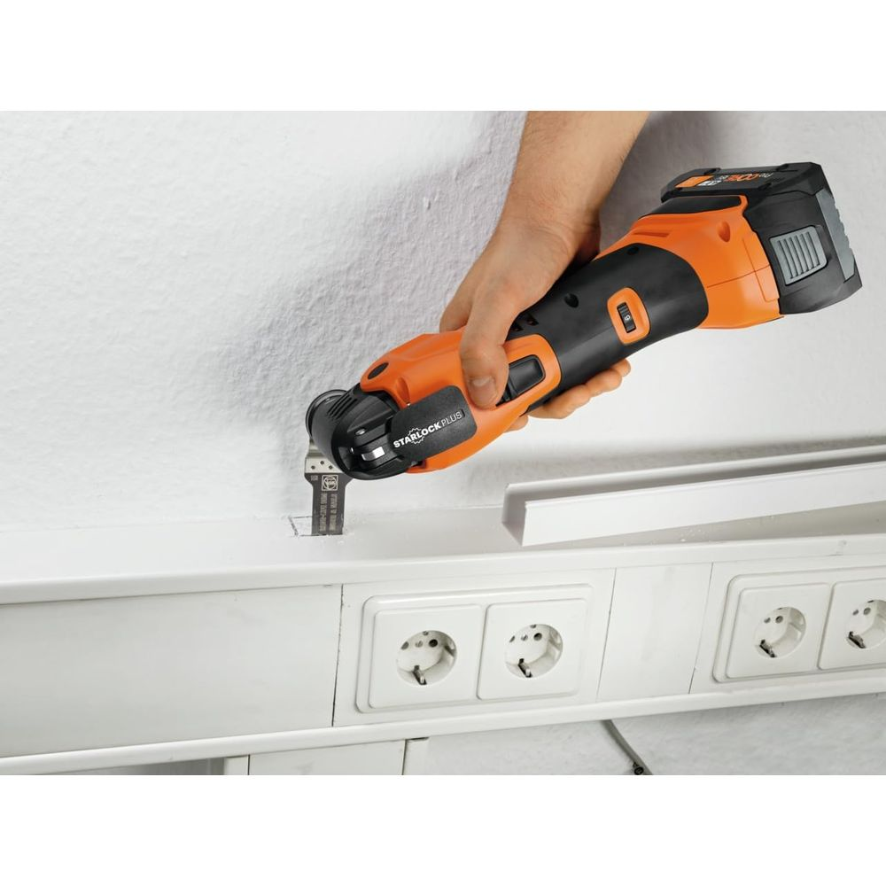 Dłoń trzyma pomarańczowo-czarne narzędzie wielofunkcyjne, tnąc białą listwę nad gniazdkami.