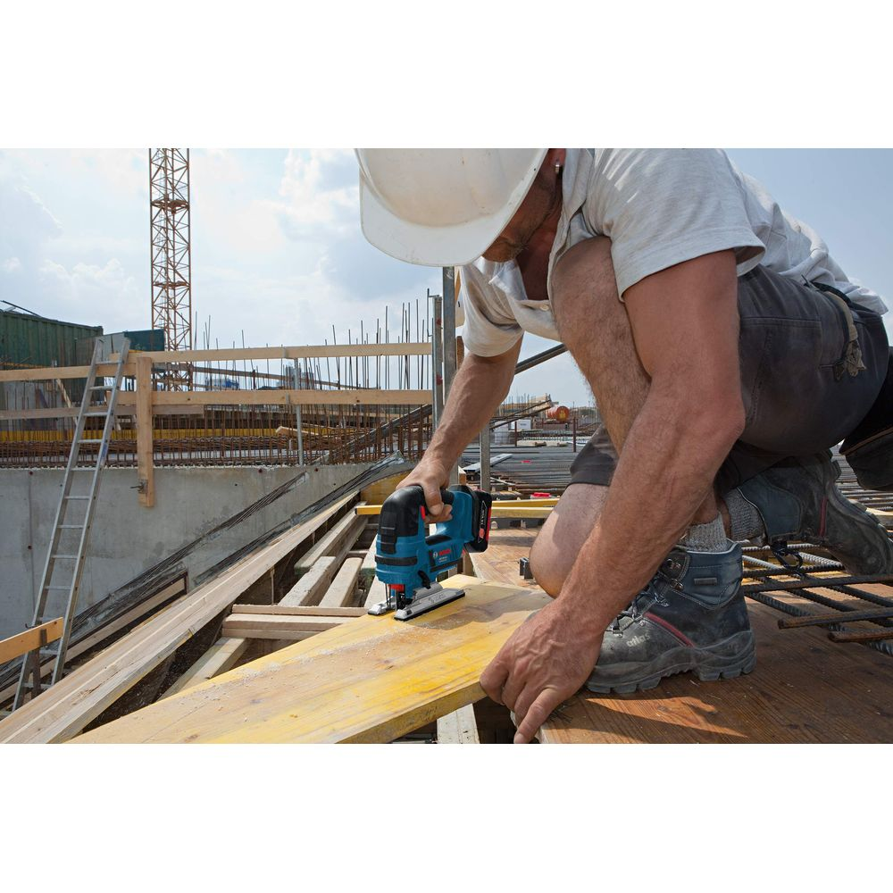 Budowlaniec tnie drewno wyrzynarką na budowie, dźwig w tle.