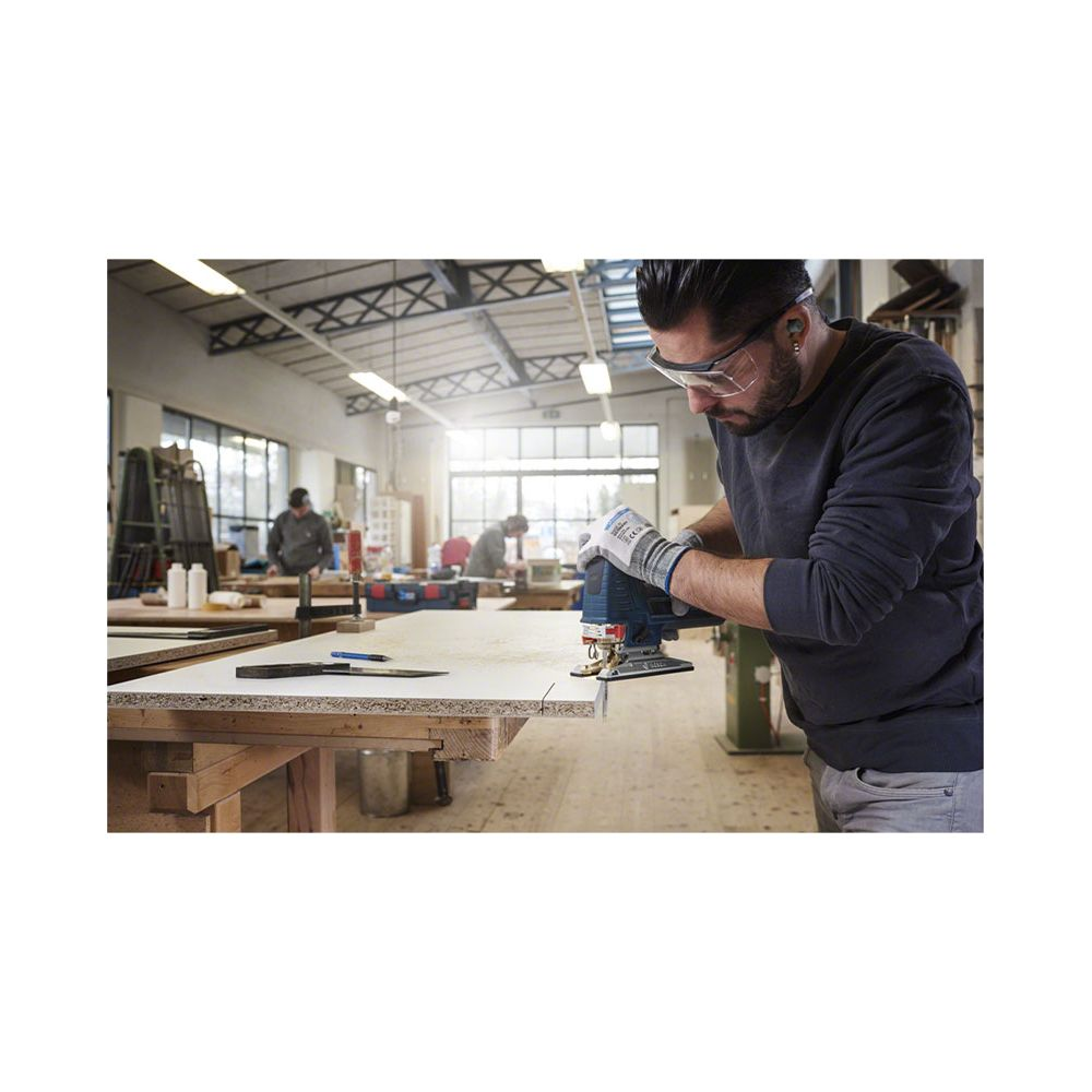 Mann mit Schutzbrille und Handschuhen schneidet Holz mit Stichsäge in Werkstatt.
