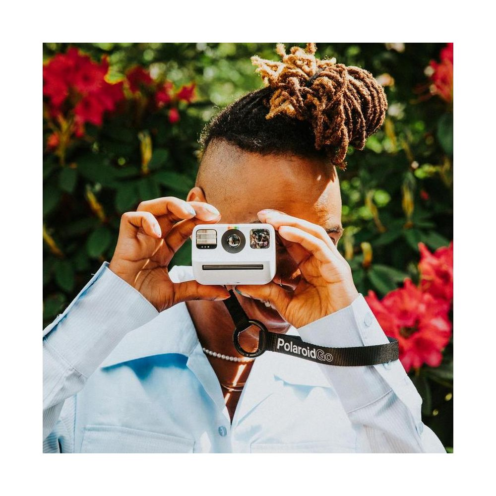 Osoba z dredami robi zdjęcie białym aparatem natychmiastowym, czerwone kwiaty w tle.