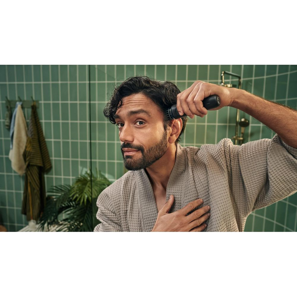 Mężczyzna z brodą i szlafrokiem używa trymera do włosów na głowie przed zielonymi kafelkami.