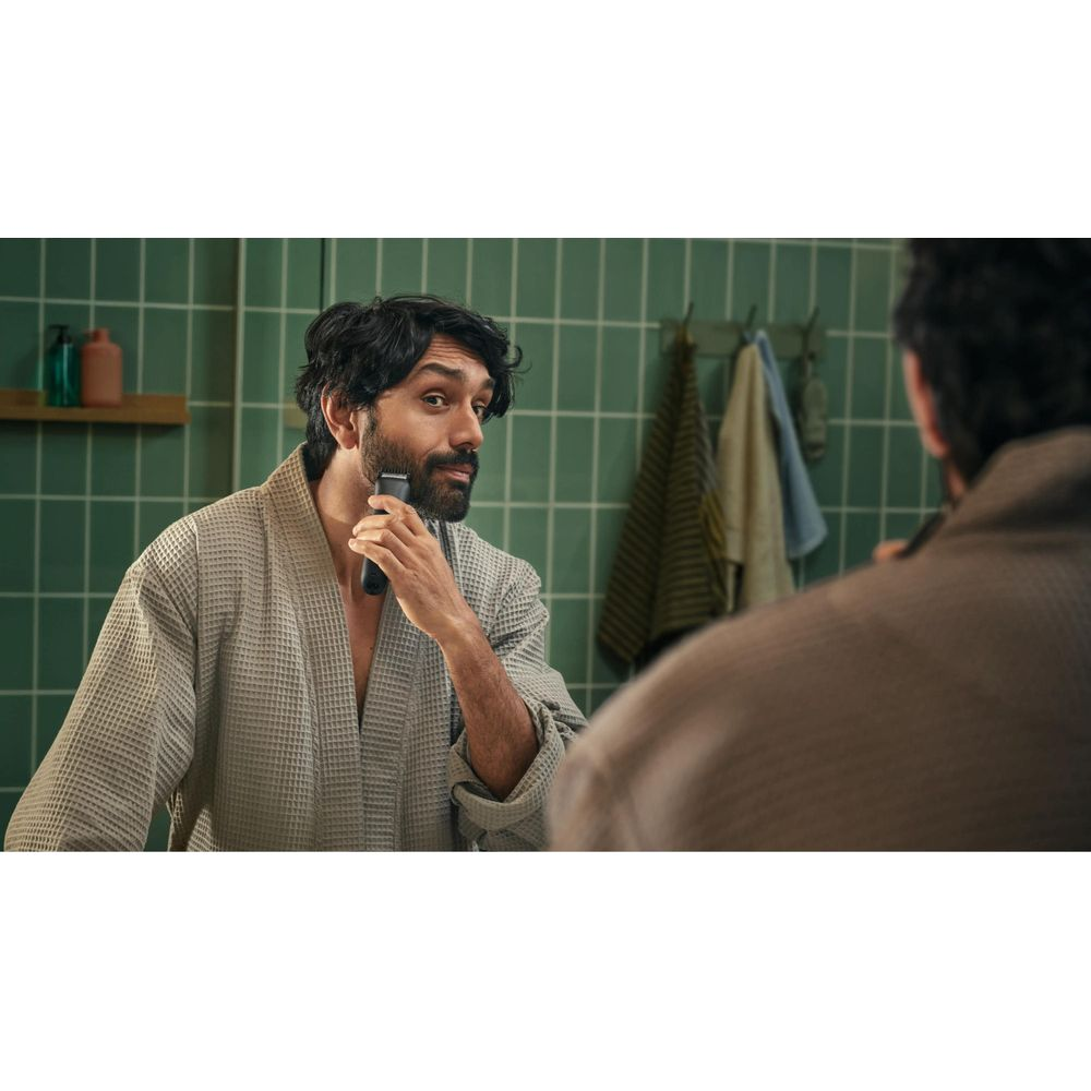 Mężczyzna z trymerem do brody w łazience, w szlafroku, przed zielonymi płytkami.