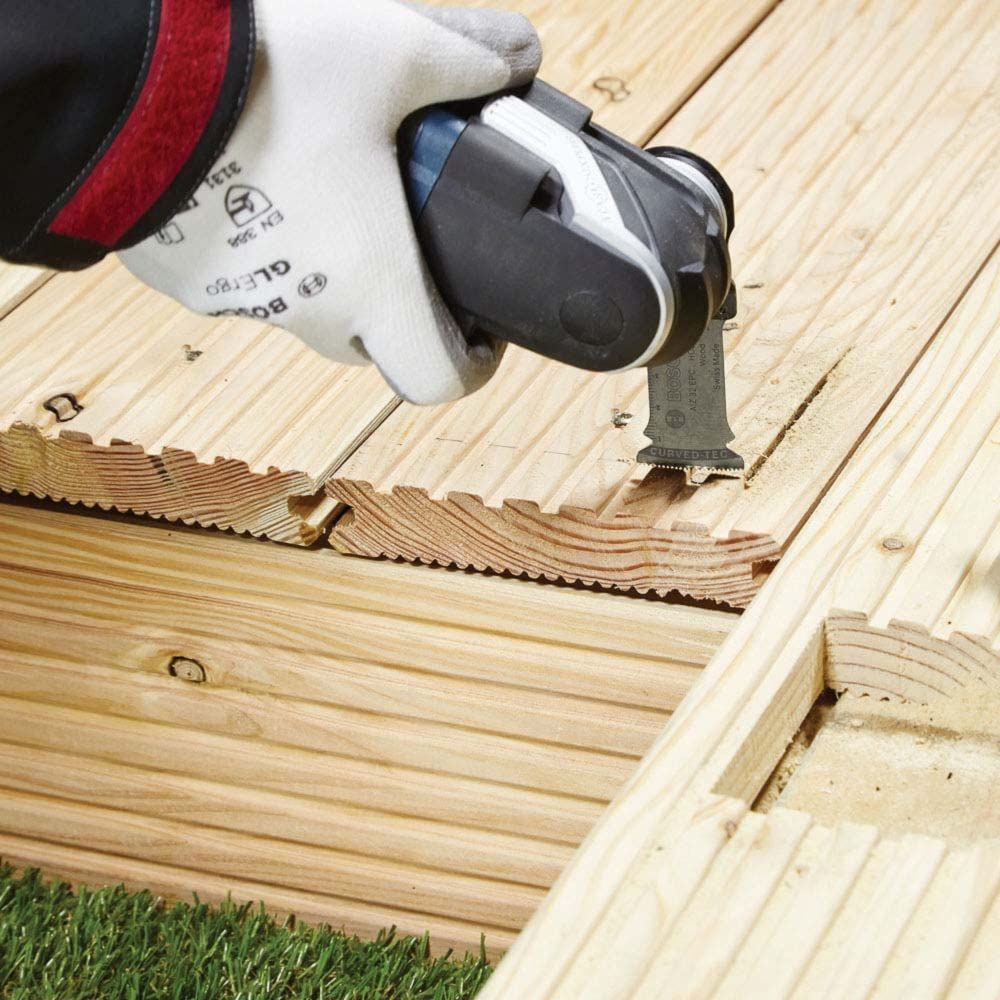 Hand mit Handschuh benutzt Oszillierwerkzeug auf Holzplanken, Gras im Vordergrund.