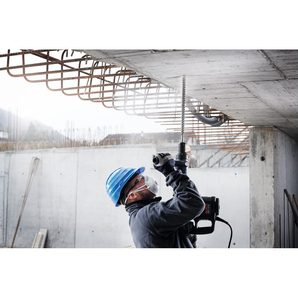 Robotnik budowlany wierci w suficie. Niebieski kask, biała maska, okulary ochronne. Widoczna stal.