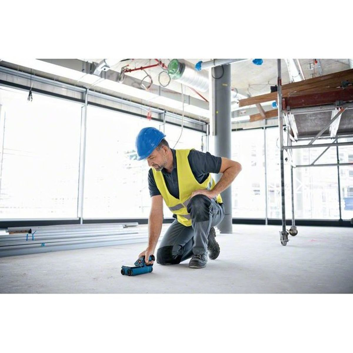 Uomo con elmetto e gilet in ginocchio usa strumento di misura in cantiere.