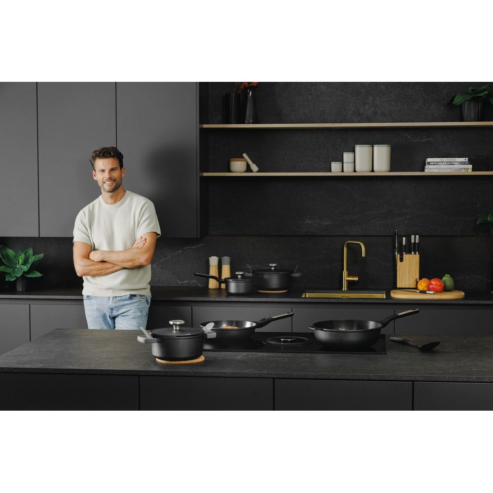 Man in casual clothes stands in a modern black kitchen with cookware.