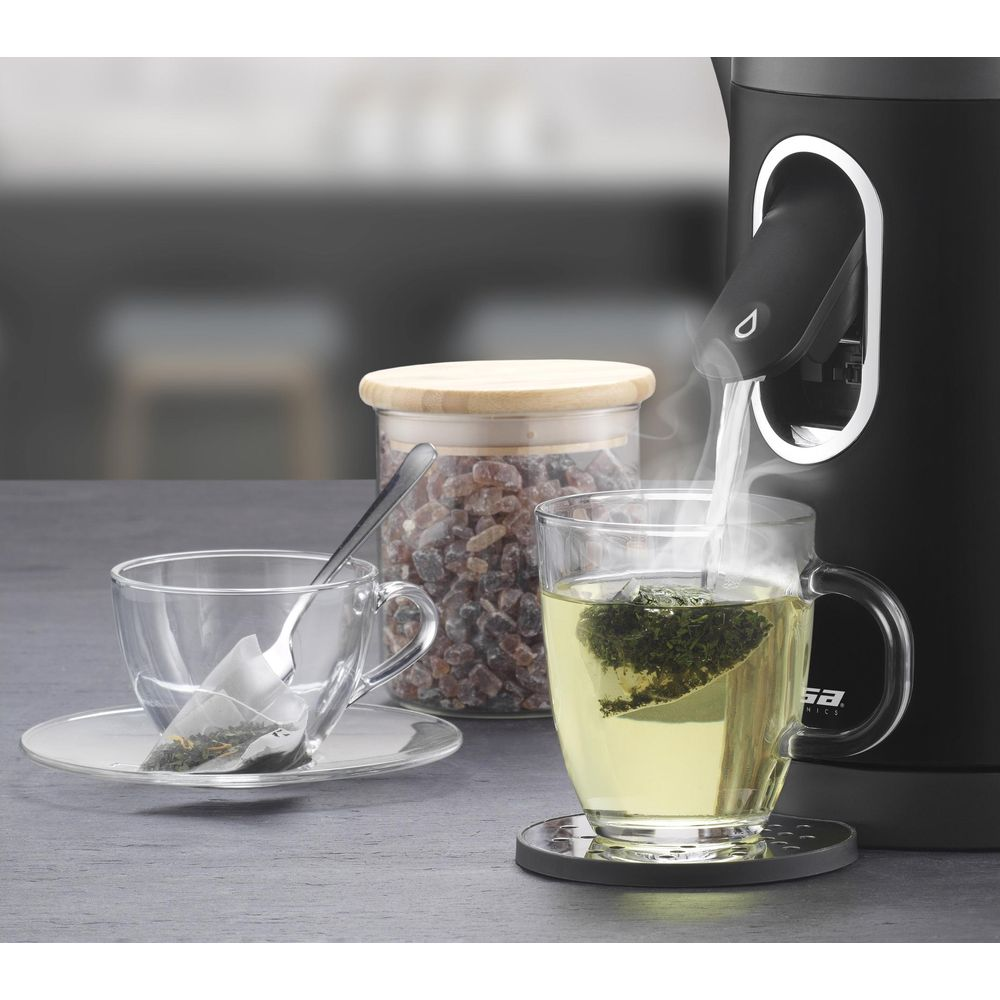 Heißes Wasser fließt aus einem schwarzen Wasserkocher in eine Teetasse. Ein Zuckerglas steht daneben.