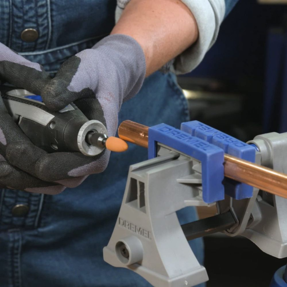 Persona con guantes usa herramienta rotatoria en tubo de cobre en soporte Dremel.