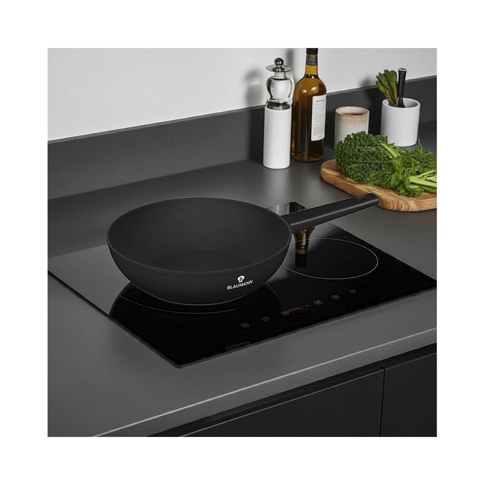 Un wok negro sobre una placa de inducción con verduras y botellas al fondo.