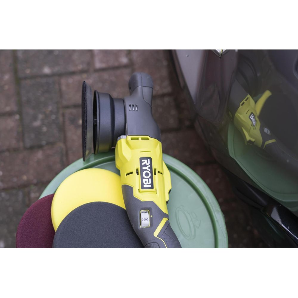 Ein gelber Ryobi Polierer mit Aufsätzen liegt auf einem grünen Eimer neben einem Auto.