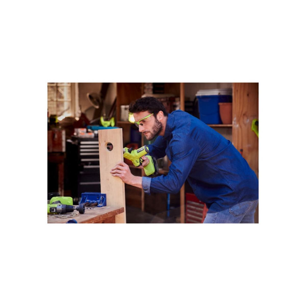 Hombre taladrando madera con herramienta verde, usa gafas y ropa de trabajo azul.