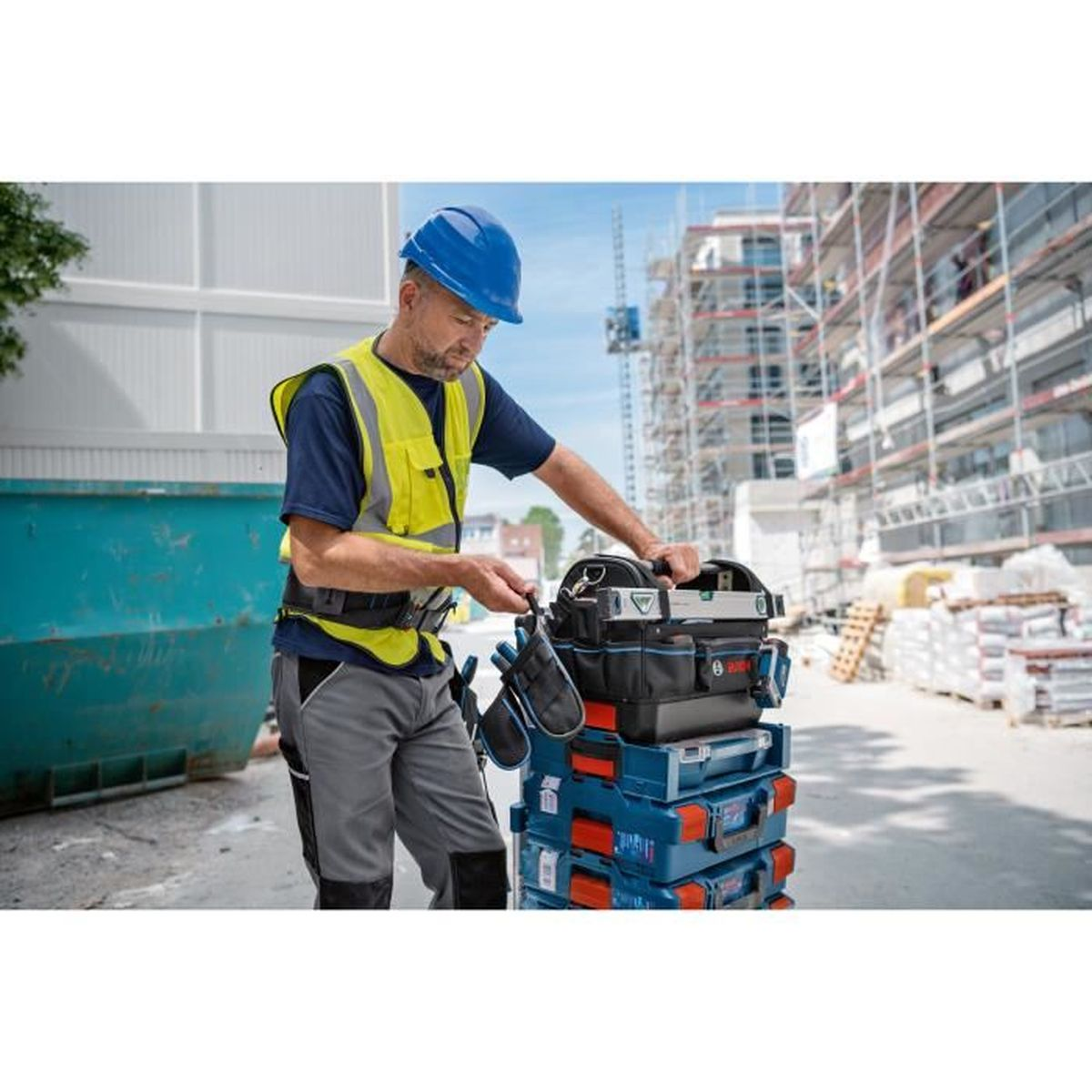 Ein Bauarbeiter in Warnweste und Helm sortiert Werkzeugkisten auf einer Baustelle.