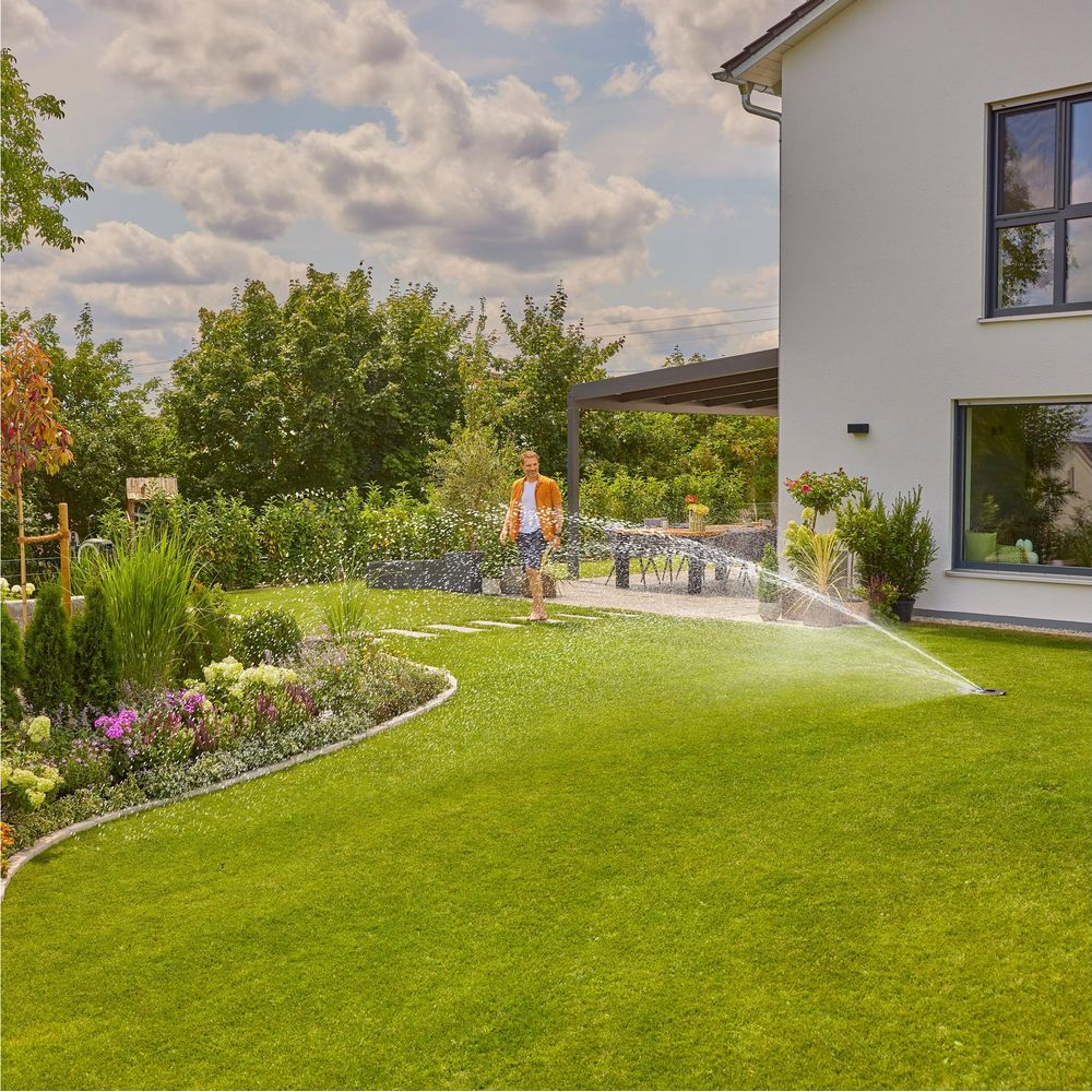 Mann bewässert Rasen vor modernem Haus mit Garten und Blumenbeeten.