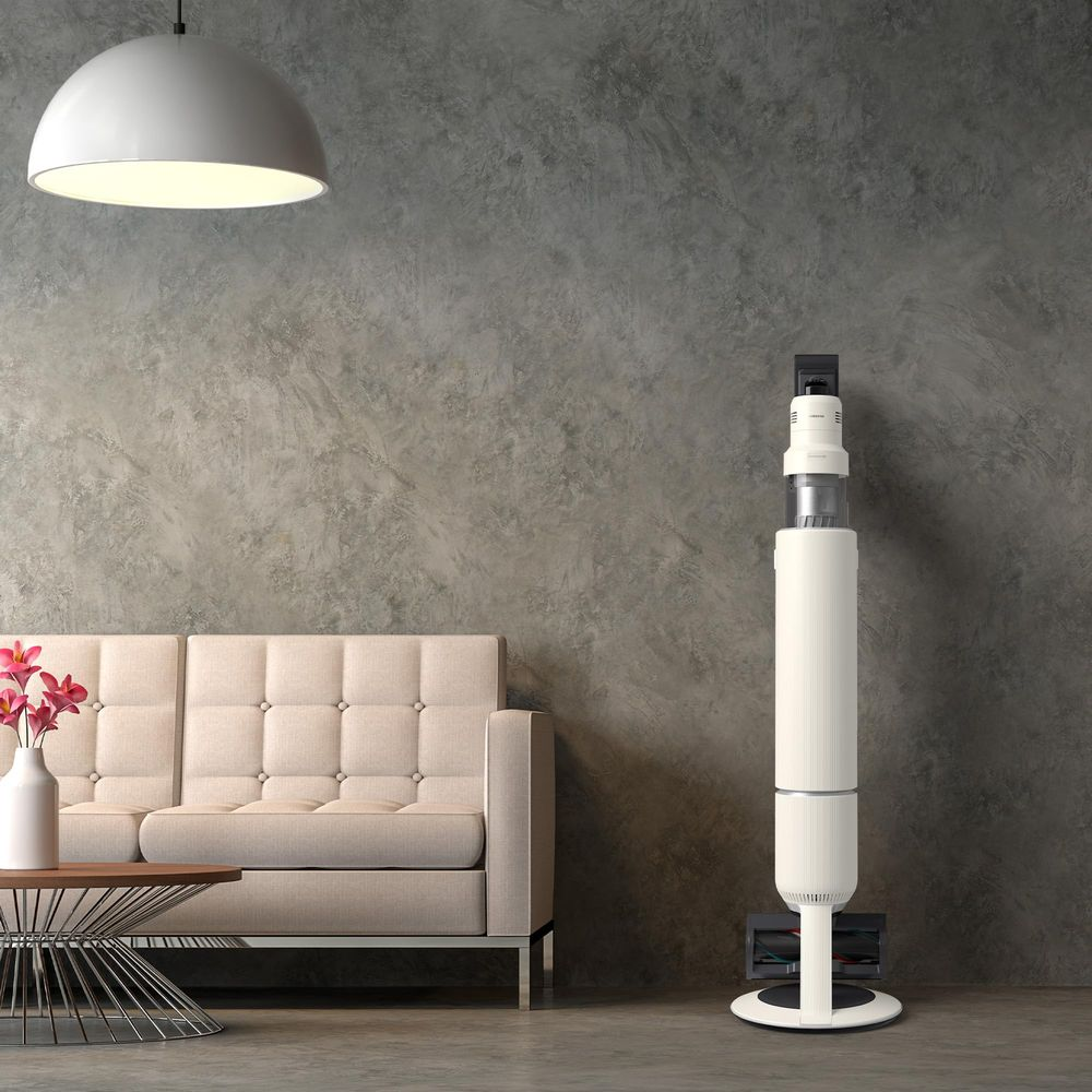 Un canapé beige près d'une table basse avec des fleurs. À droite, un aspirateur blanc.