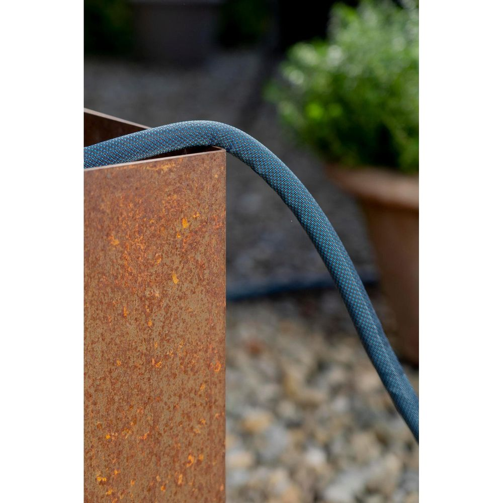 Ein blauer Gartenschlauch ruht auf dem Rand eines rostigen Metallpflanzgefäßes, mit unscharfem Grün im Hintergrund.