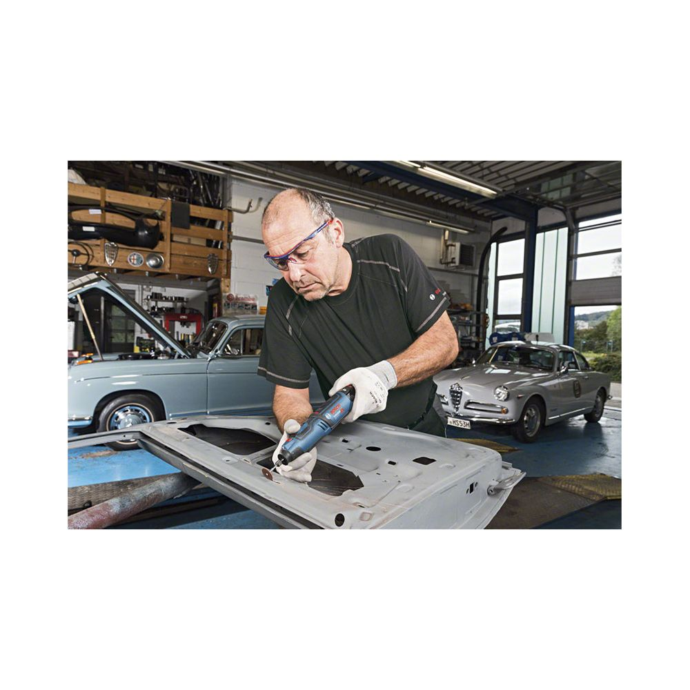 Mężczyzna w warsztacie naprawia drzwi samochodu narzędziem, stare samochody w tle.