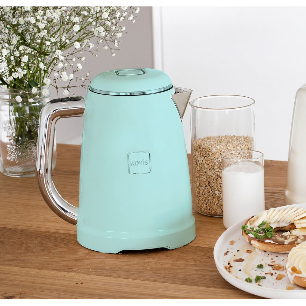 Ein tealfarbener Novis-Wasserkocher steht auf einem Holztisch mit Haferflocken, Milch und einem Bagel.