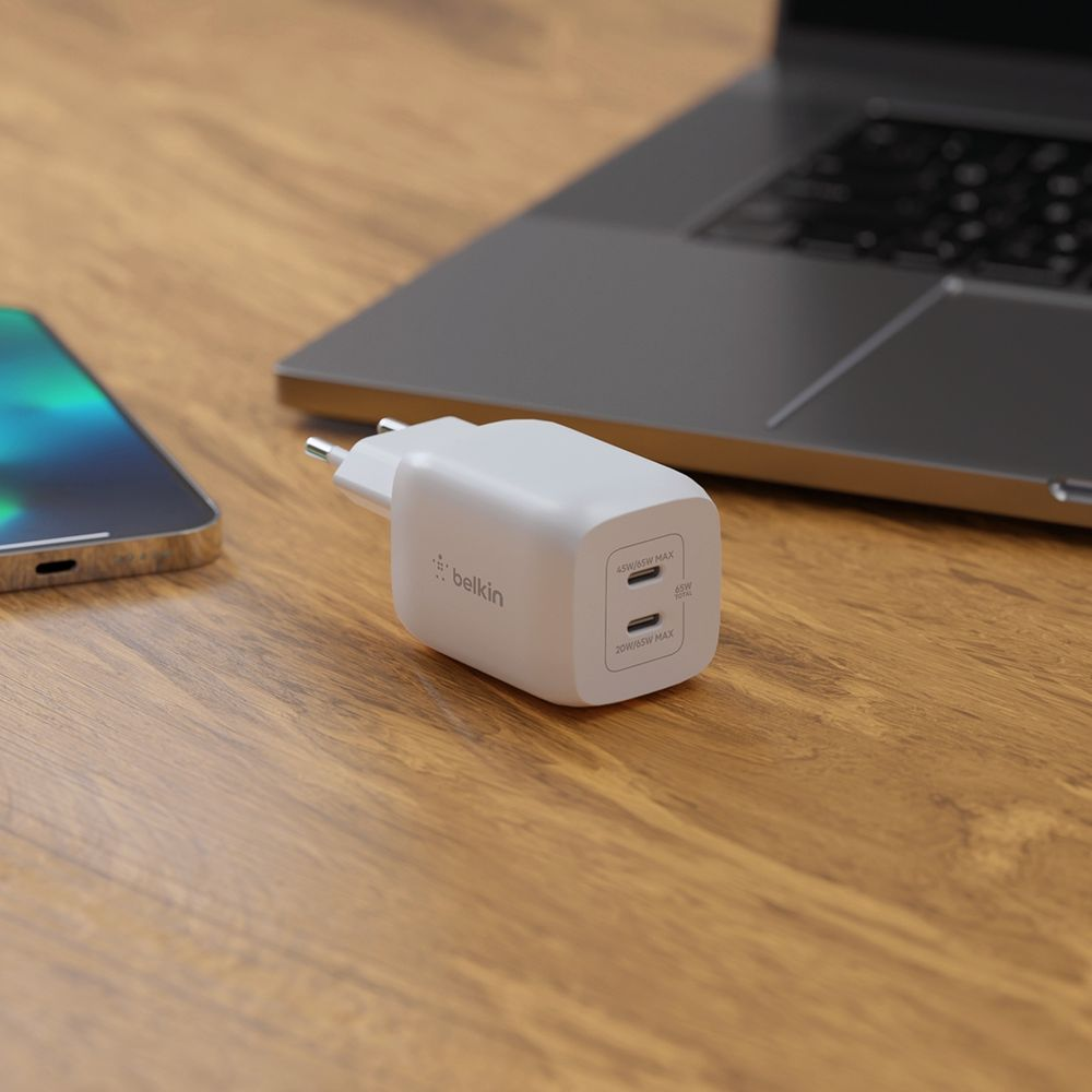 Un caricatore Belkin USB-C bianco a doppia porta su un tavolo di legno accanto a uno smartphone e un laptop.