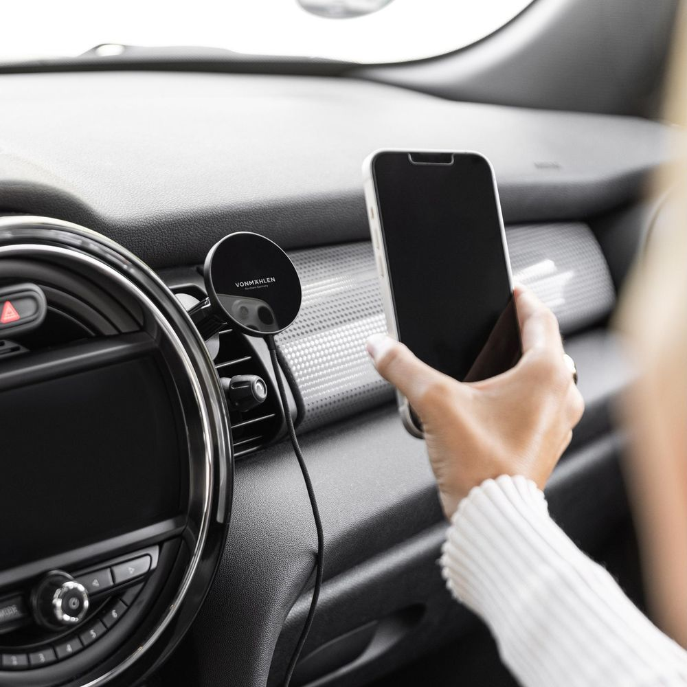 Hand hält Handy am Armaturenbrett neben Lenkrad und Lüftungsschlitz.