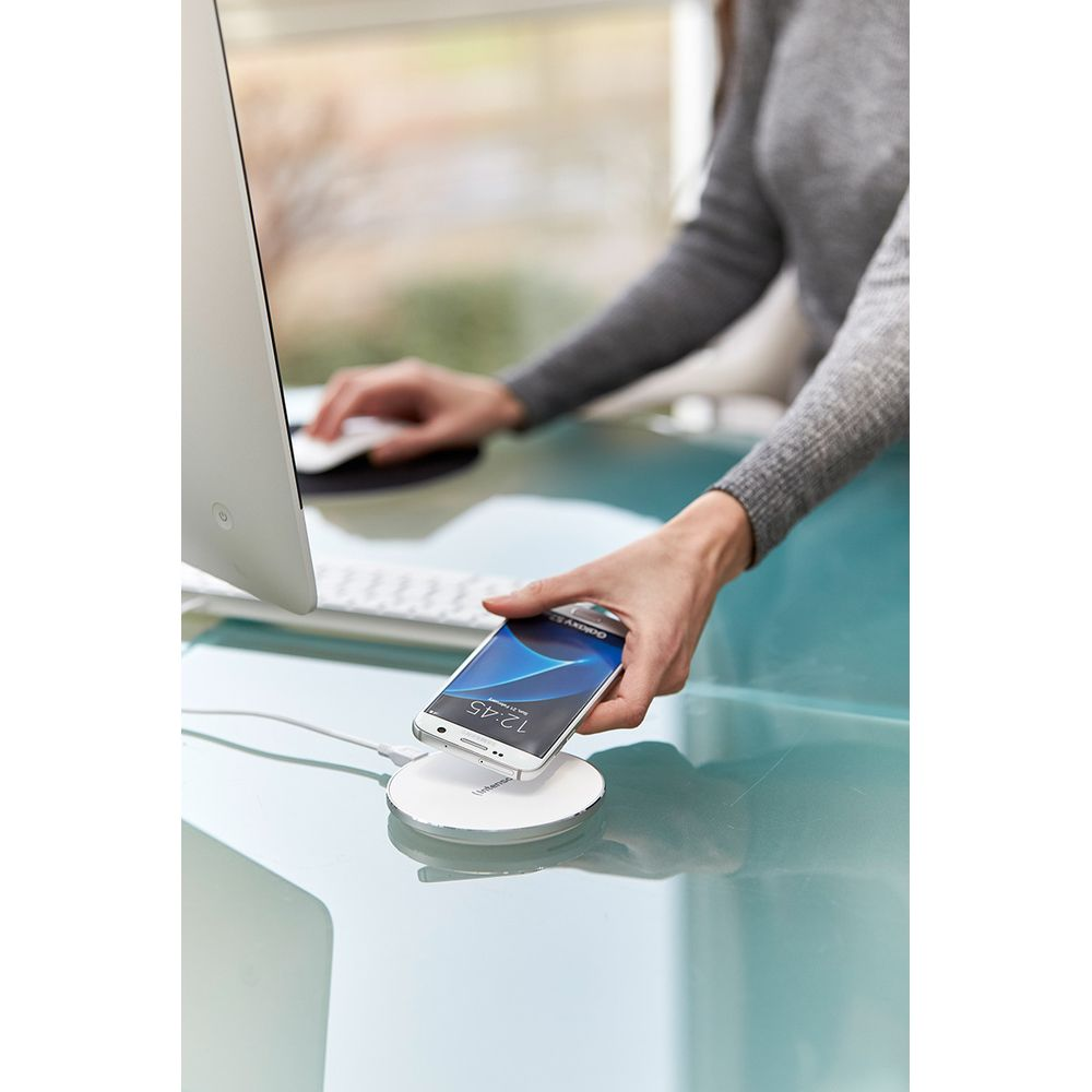 Eine Hand legt ein Smartphone auf ein weißes kabelloses Ladegerät neben einem Computer.