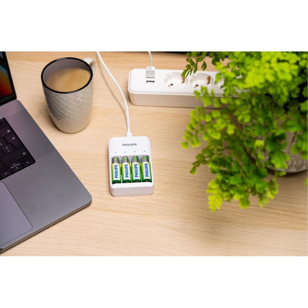 Ein weißes Philips Ladegerät mit vier AA-Batterien und ein Laptop auf einem Holztisch.