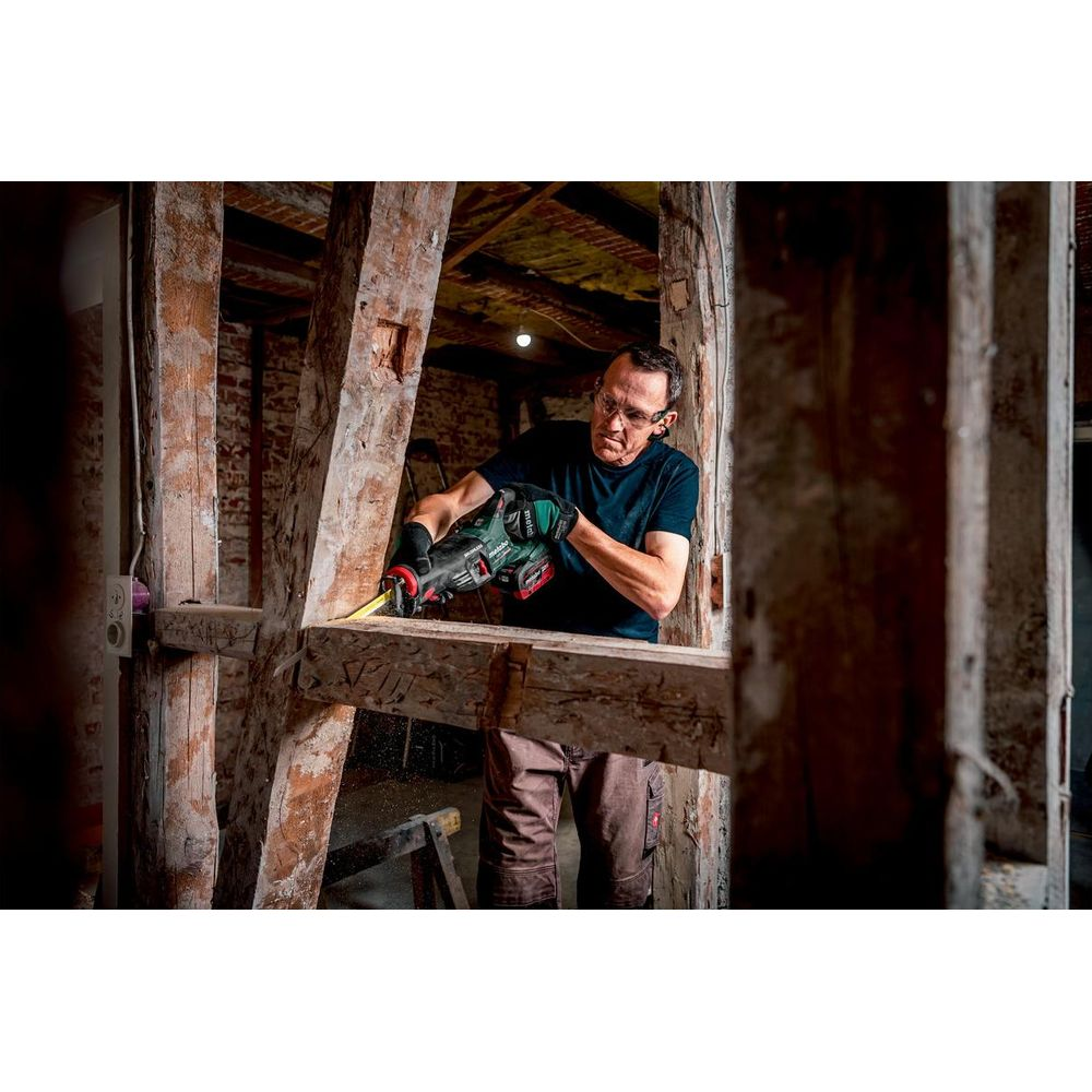 Mężczyzna w okularach ochronnych i rękawicach piłuje drewno piłą szablastą w rustykalnym otoczeniu.