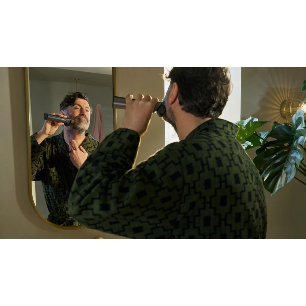 Uomo si rade allo specchio con rifinitore per barba, indossa accappatoio verde.