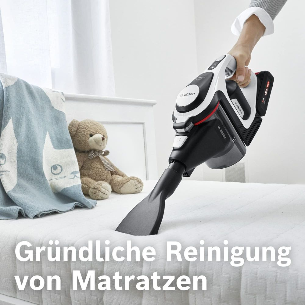 Ein Handstaubsauger reinigt eine Matratze, neben einem Teddybär und einer Decke.