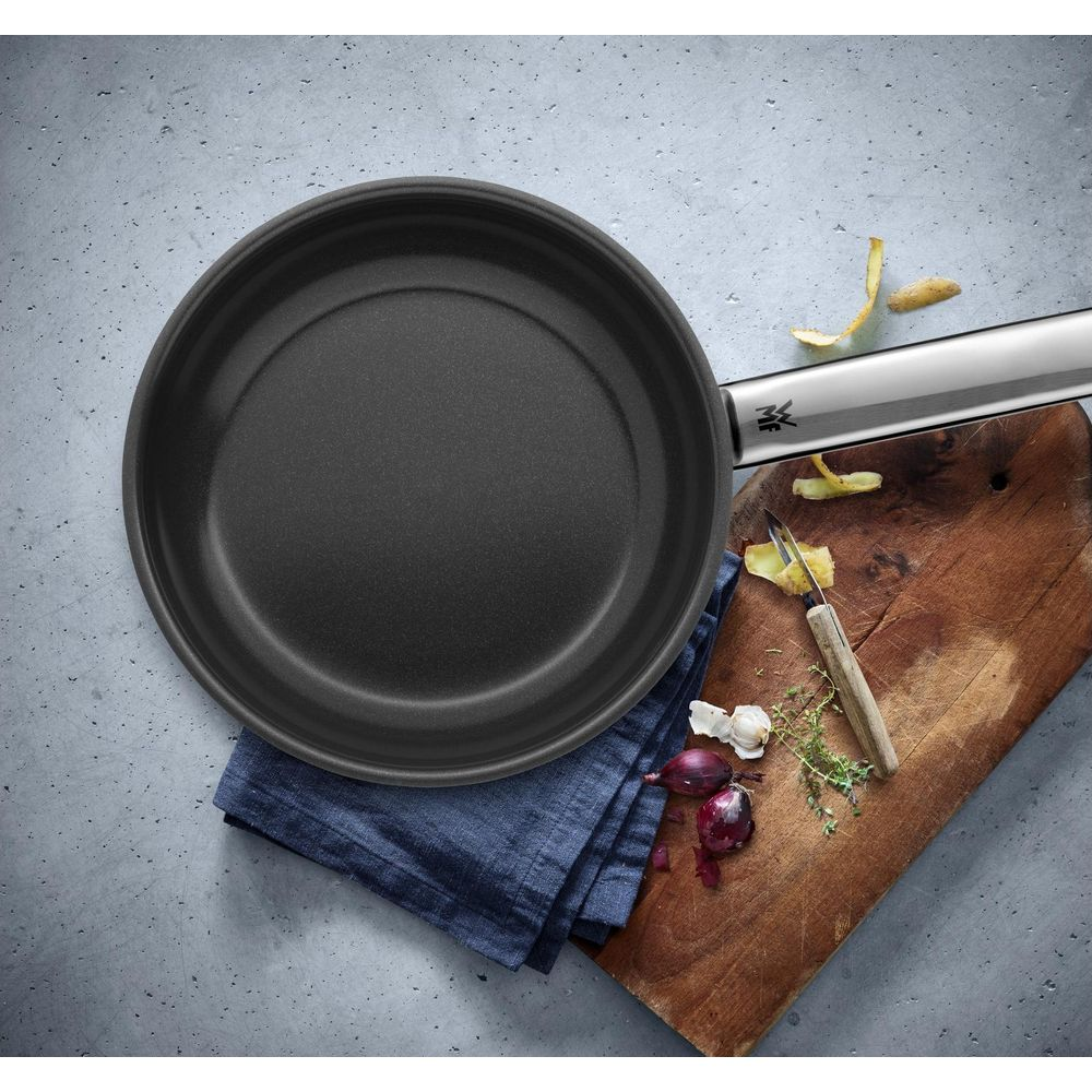 Una padella nera poggia su un panno blu accanto a cipolle e un pelapatate.