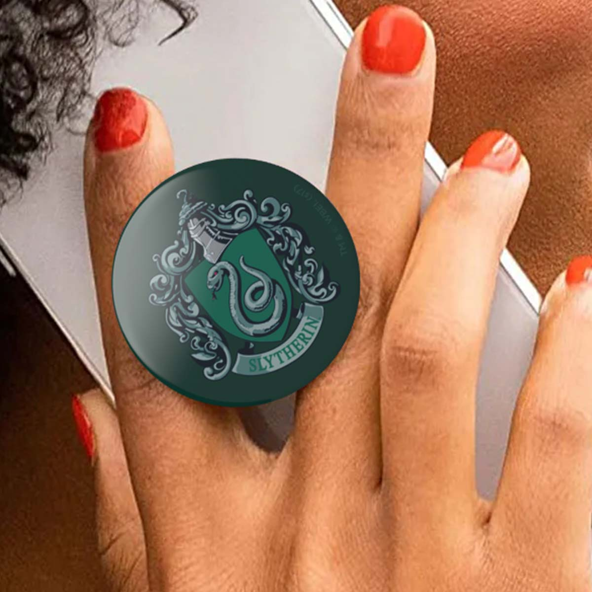 Escudo verde de Slytherin con serpiente en un agarre de teléfono en dedos con esmalte rojo.