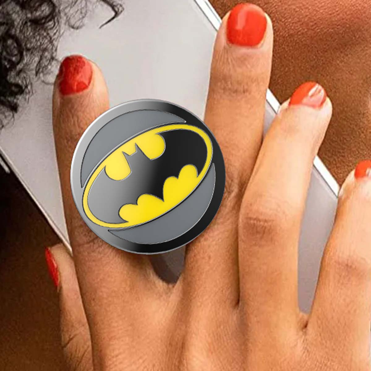 Una mano con uñas rojas sostiene un smartphone. Un logo de Batman está en un agarre.