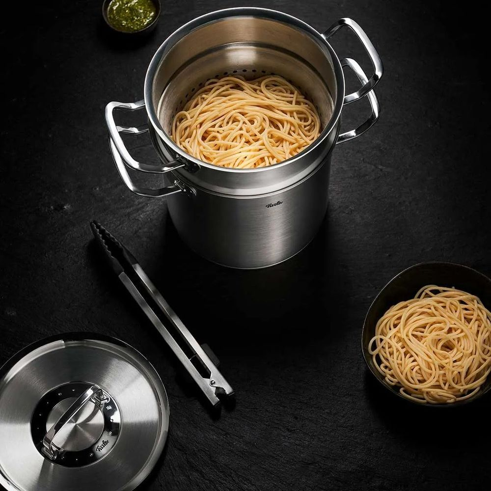 Una pentola in acciaio con spaghetti, accanto una ciotola di pesto e pinze.