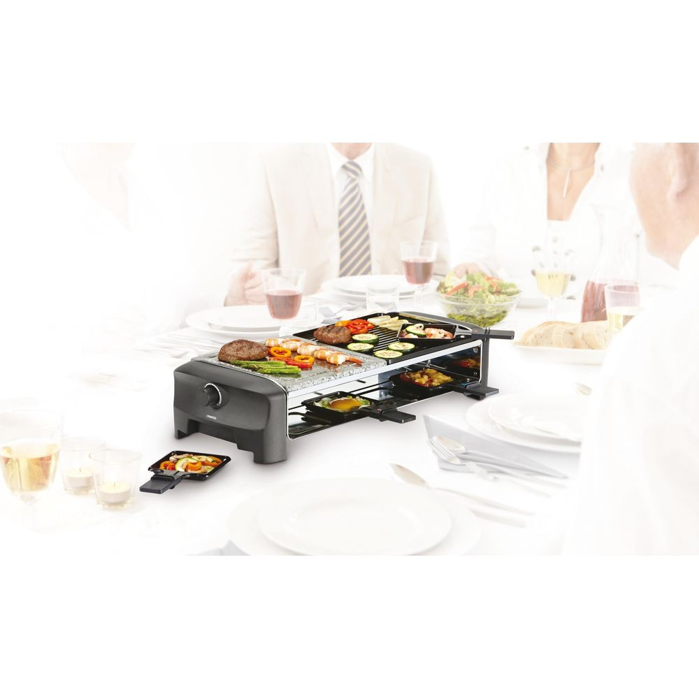 Ein Raclette-Grill mit Fleisch und Gemüse steht auf einem Tisch. Leute essen.