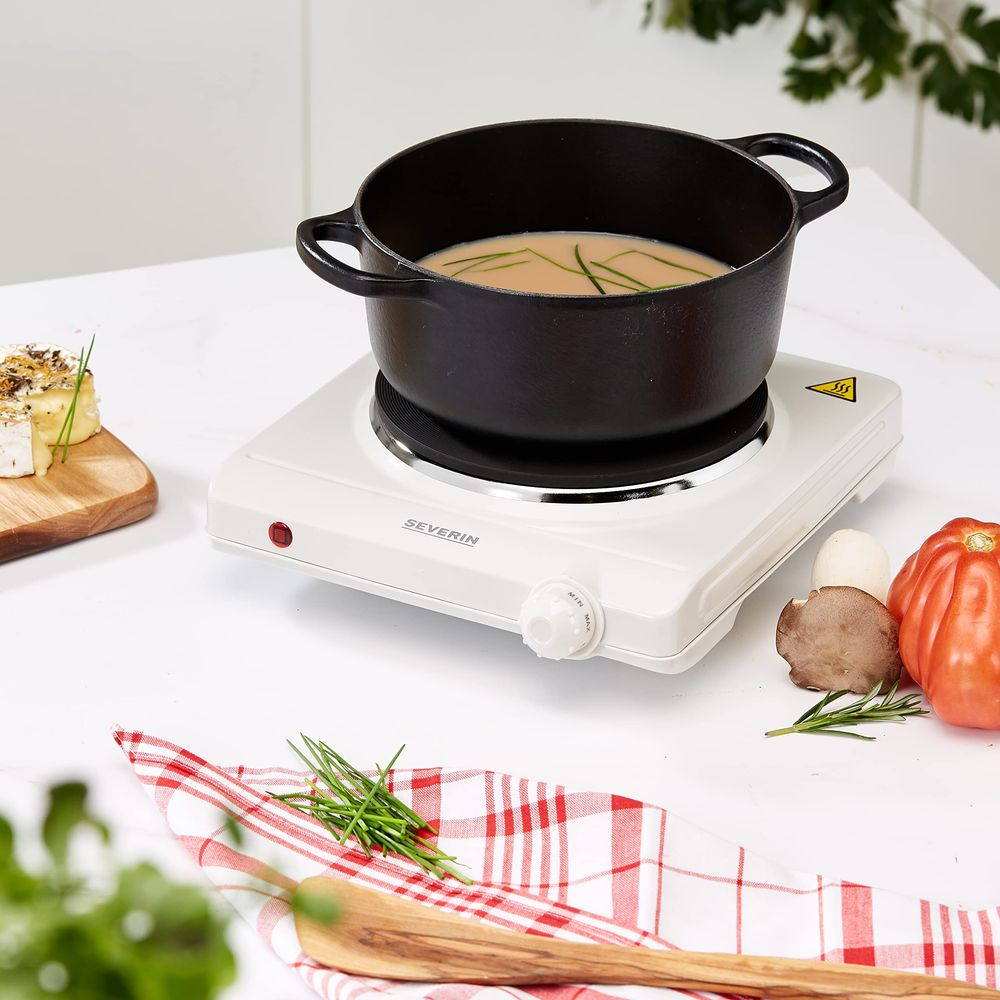 Garnek z zupą stoi na białej płycie grzewczej obok sera i warzyw.