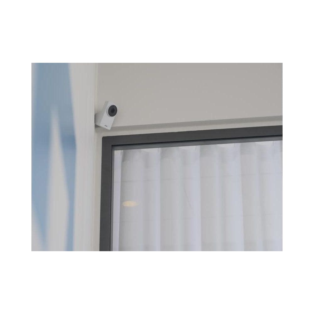 Mała biała kamera jest zamontowana na jasnoszarym murze nad oknem z białymi zasłonami.