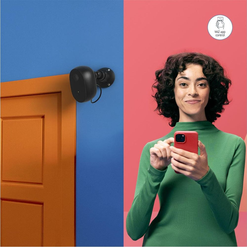 A black security camera mounted on a blue wall next to an orange door. A woman holds a red phone.