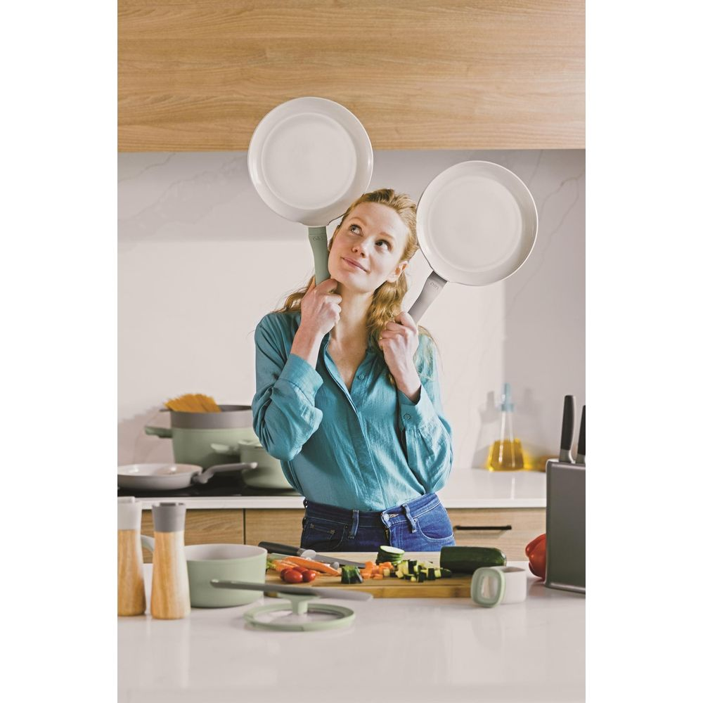A woman holds two pans over her head, with kitchenware in the foreground.
