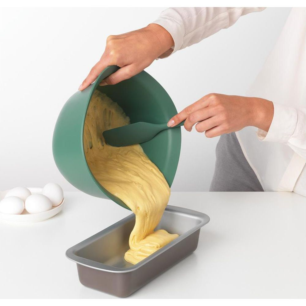 Ciasto wylewane z zielonej miski do formy keksowej. Jajka po lewej.