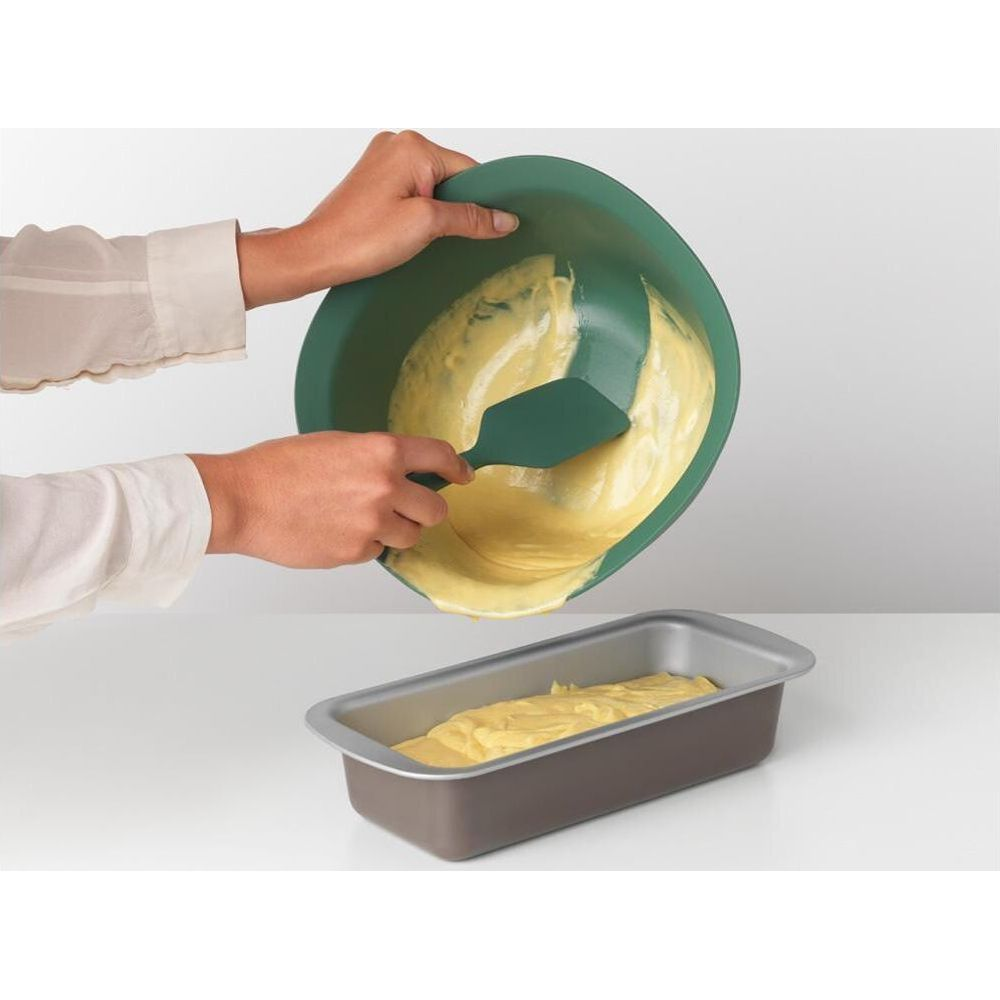 Ręce przelewają ciasto z zielonej miski do prostokątnej formy do pieczenia łopatką.
