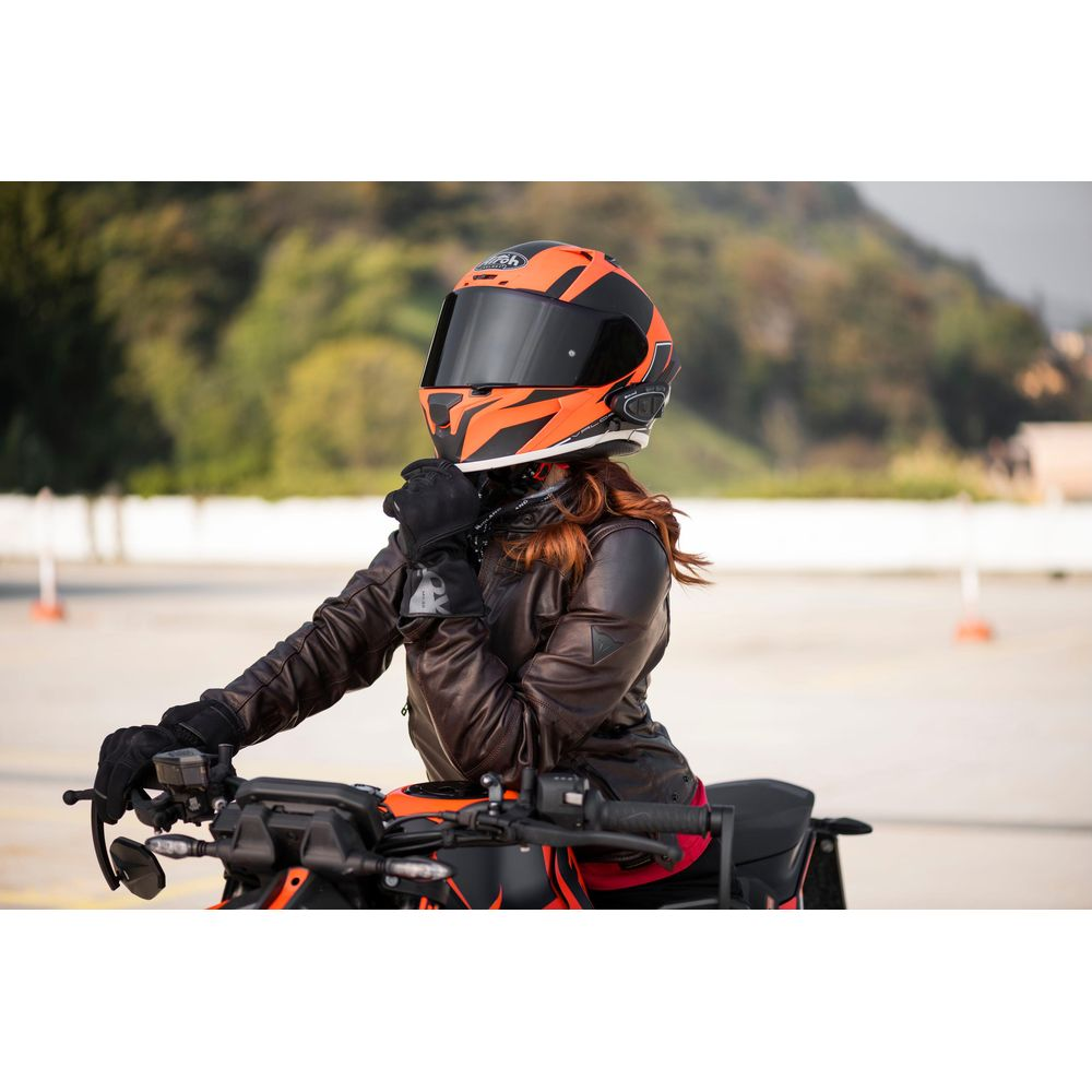 Kobieta w brązowej skórzanej kurtce i pomarańczowo-czarnym kasku siedzi na motocyklu.