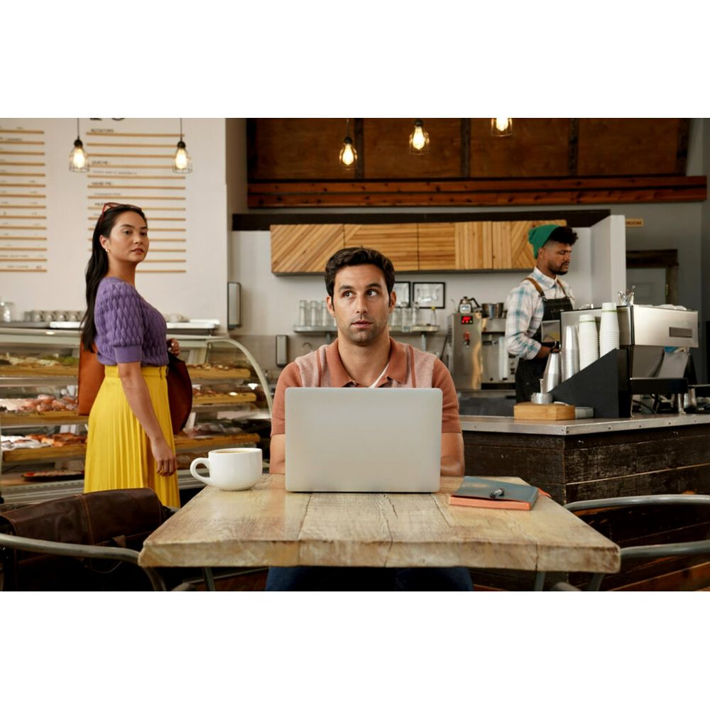 Mann mit Laptop am Tisch in einem Café, Frau und Barista im Hintergrund.