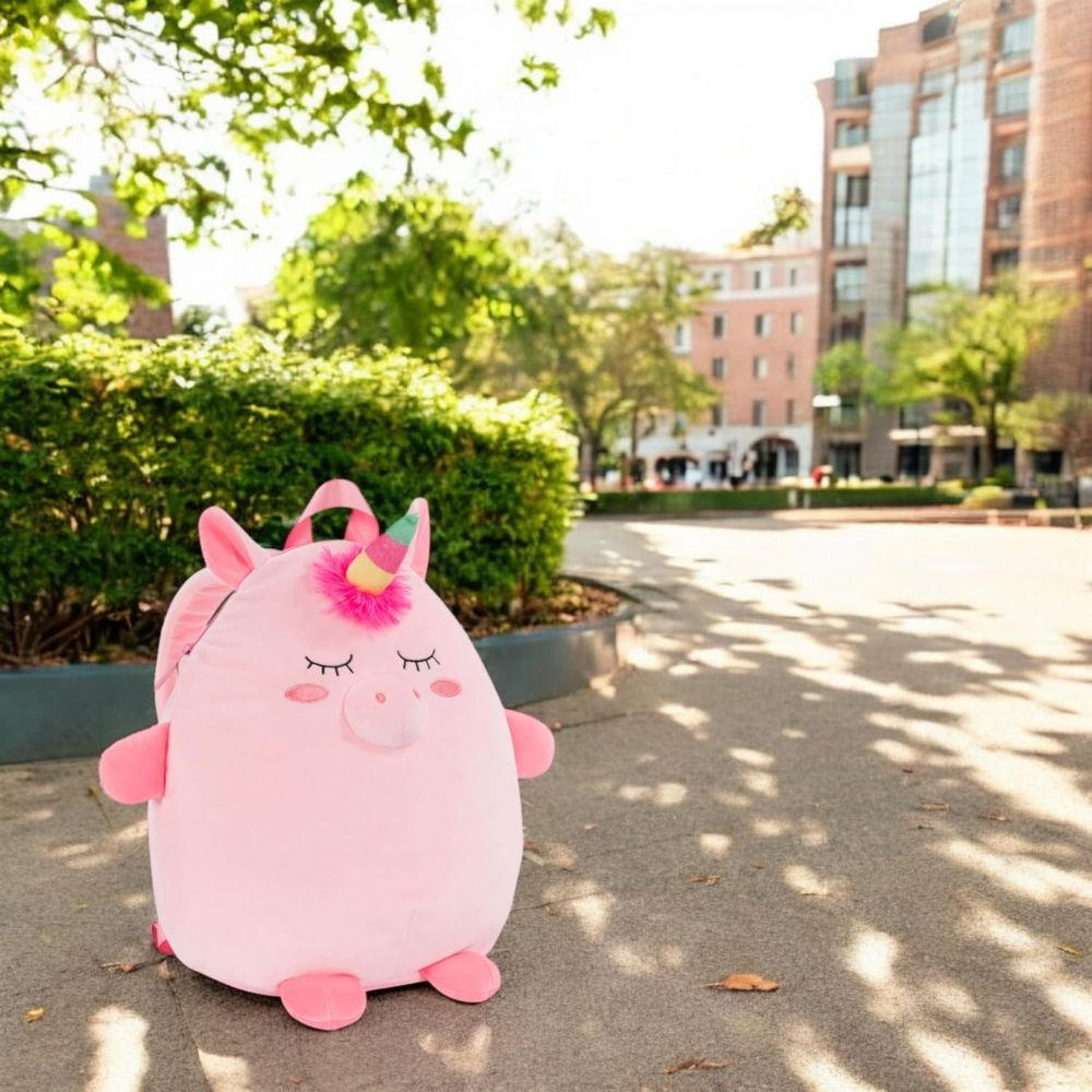 Ein rosa Plüsch-Einhornrucksack sitzt auf einem gepflasterten Weg mit Bäumen und Gebäuden im Hintergrund.